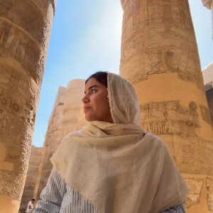 Retrato de una mujer con pañoleta color crema sobre la cabeza y los hombros, entre columnas de piedra talladas con jeroglíficos de un templo egipcio.